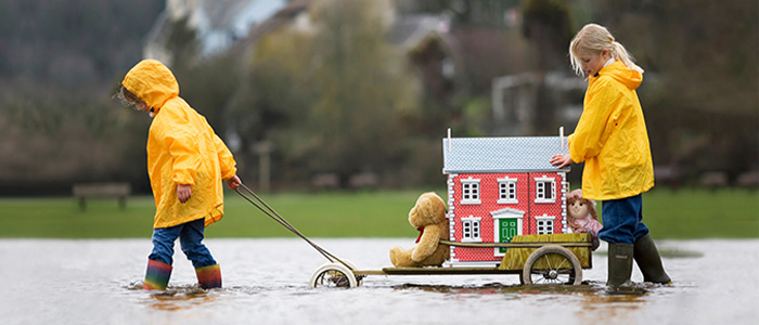 Zwei Kinder in gelben Regenjacken ziehen einen Wagen mit einem Spielzeughaus und Kuscheltieren durch überflutetes Wasser.