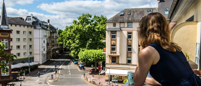 Frau blickt von einem Balkon auf eine sonnige Stadtstraße mit historischen Gebäuden.