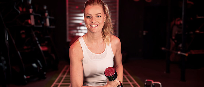 Junge Frau trainiert mit einer Hantel im Fitnessstudio und lächelt in die Kamera.