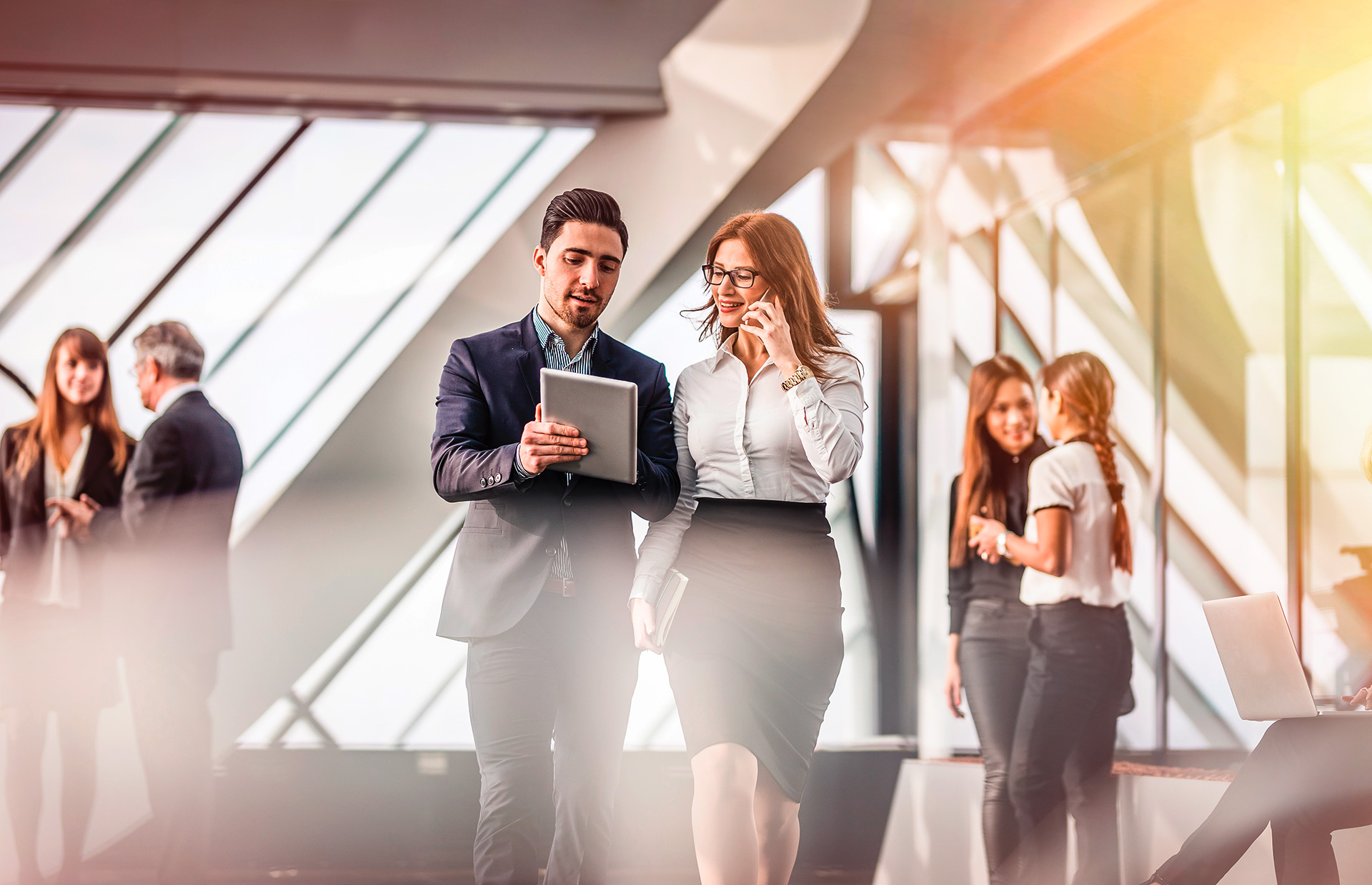 Geschäftsleute im modernen Bürogebäude, zwei Personen im Vordergrund im Gespräch und mit Tablet, weitere im Hintergrund im Austausch.