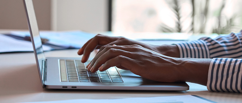 Person tippt auf einem Laptop auf einem Schreibtisch mit Dokumenten.