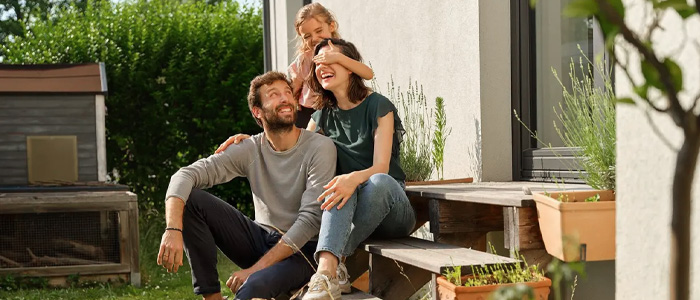Familie sitzt lachend auf der Terrasse vor ihrem Haus und genießt die Sonne.