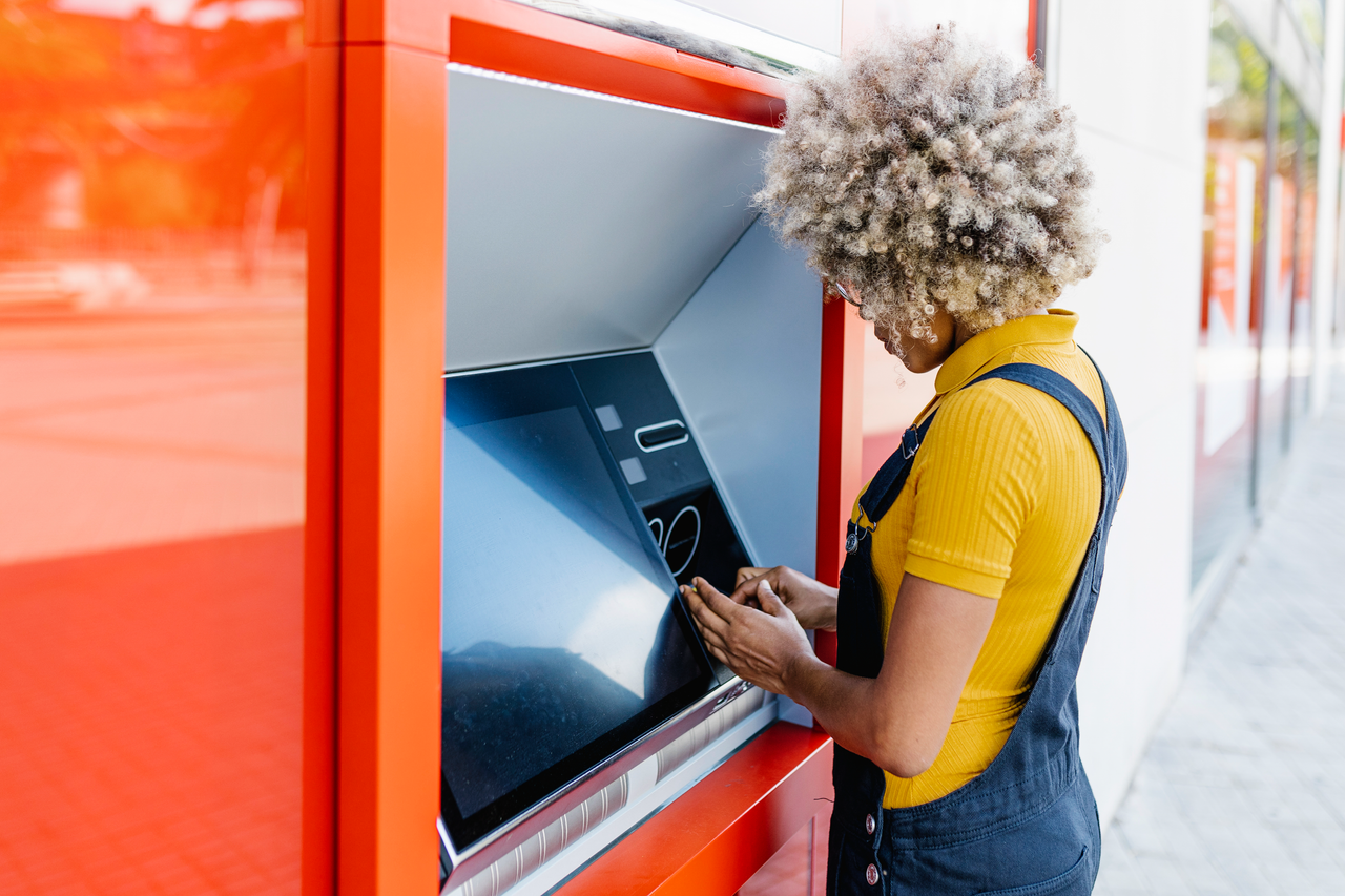 Frau steht an einem roten Geldautomaten im Freien und gibt ihre PIN ein.